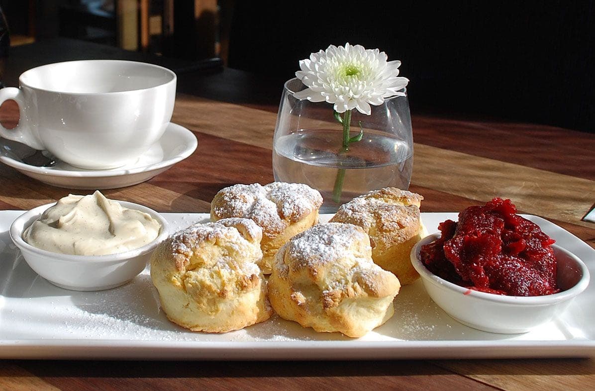 A plate of scones with jam and cream, a teacup and saucer, and a single white chrysanthemum in a glass of water on a wooden table