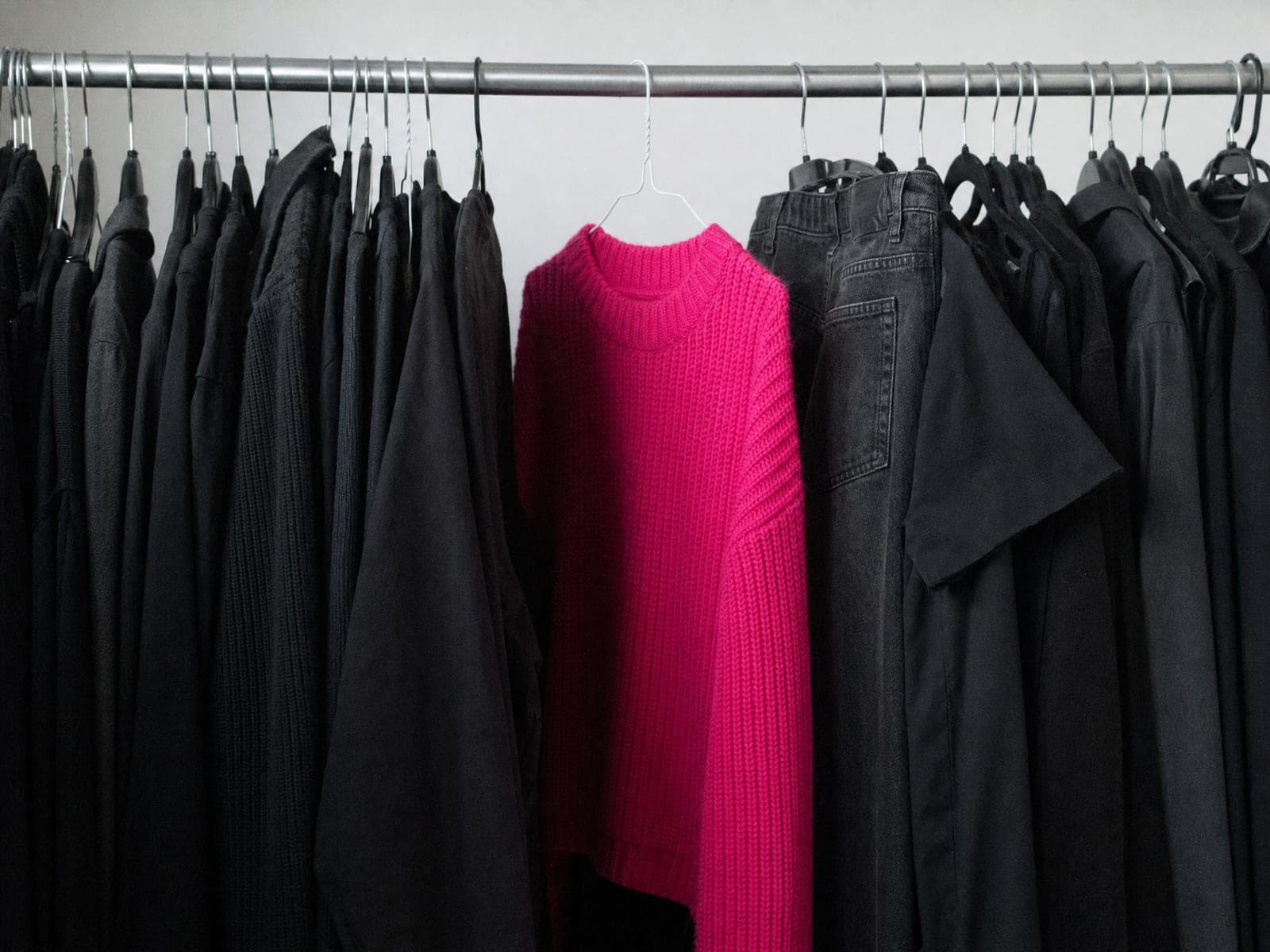 A bright fuchsia knitted jumper hanging on a clothes rail, surrounded on both sides by clothing that is entirely black and dark grey