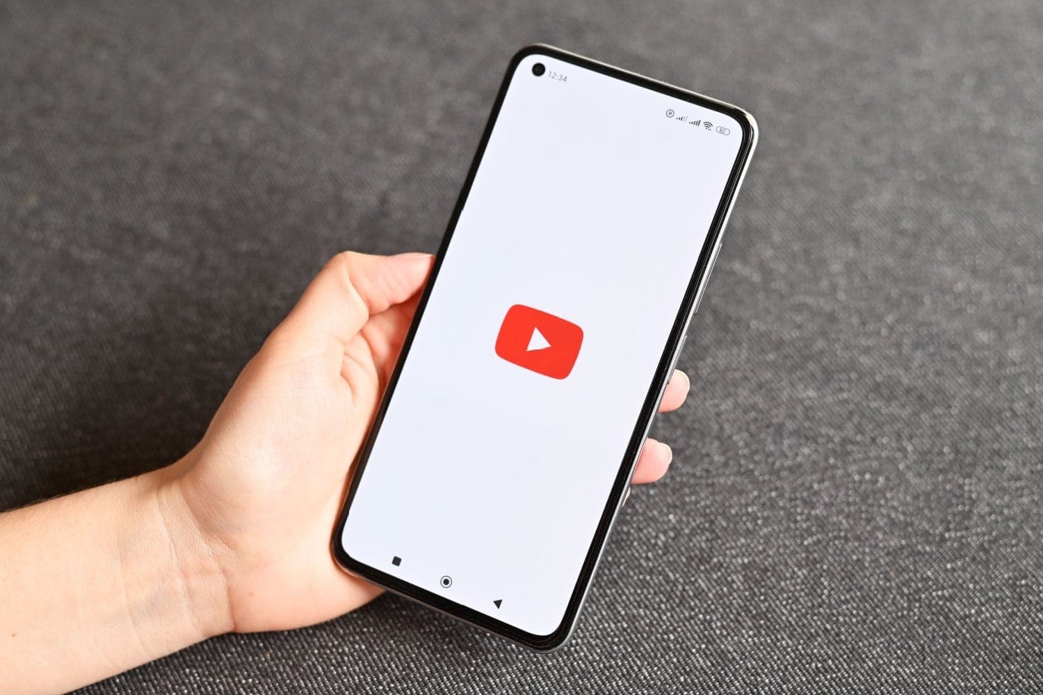 A hand holding a smartphone showing the YouTube app loading screen, with a grey fabric background.