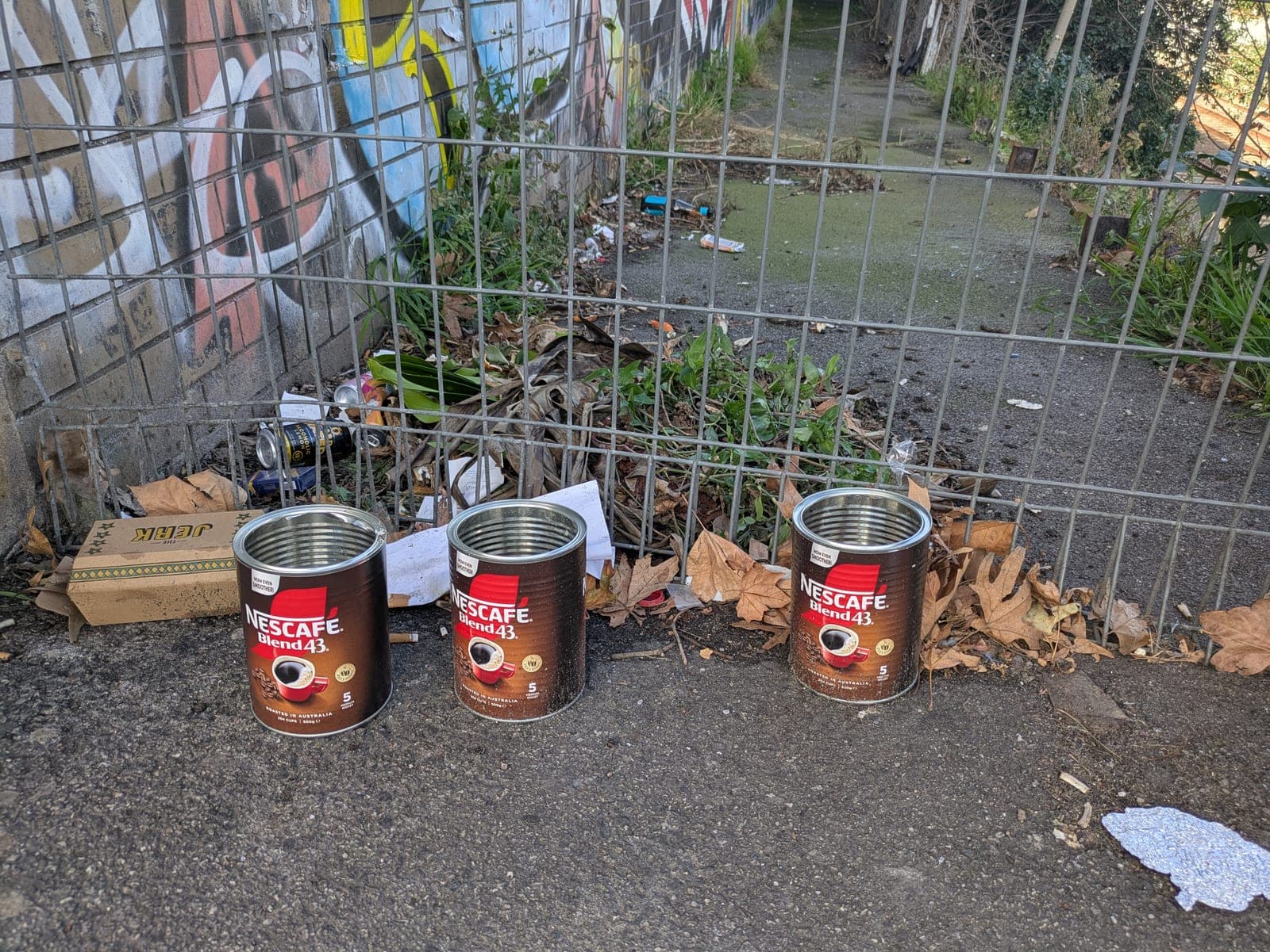Three tins of Nescafé Blend 43 lined up on a footpath beside a wire fence