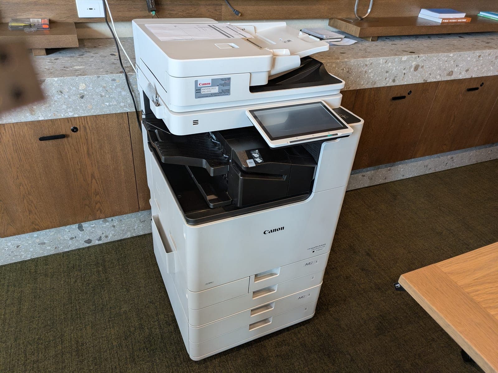A large Canon multifunction office printer standing beside a desk, with printed pages sitting in the output tray.