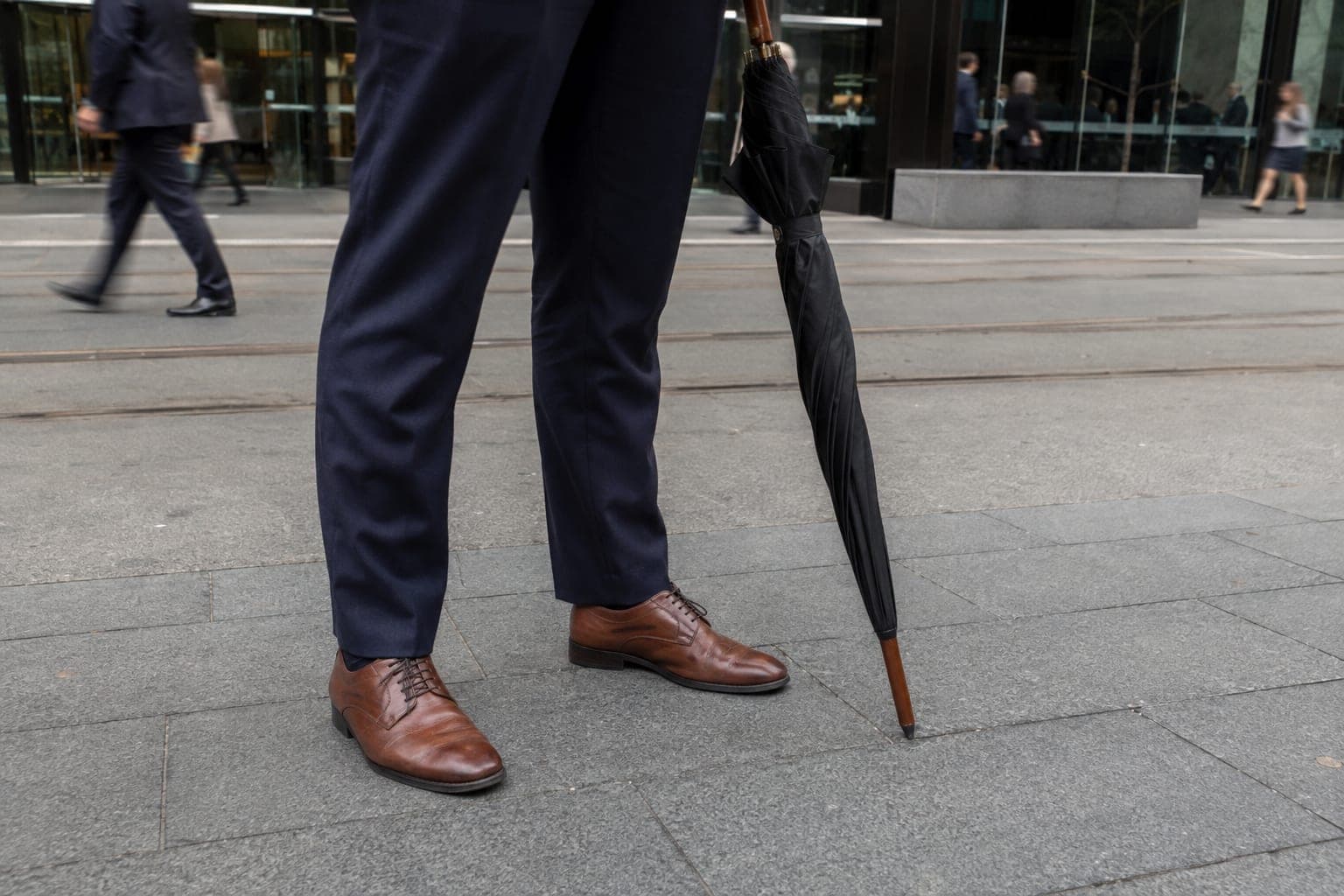 A man in office attire walking through the Melbourne CBD carrying a large, fully closed golf umbrella tip-down like a walking stick, under a clear sky.