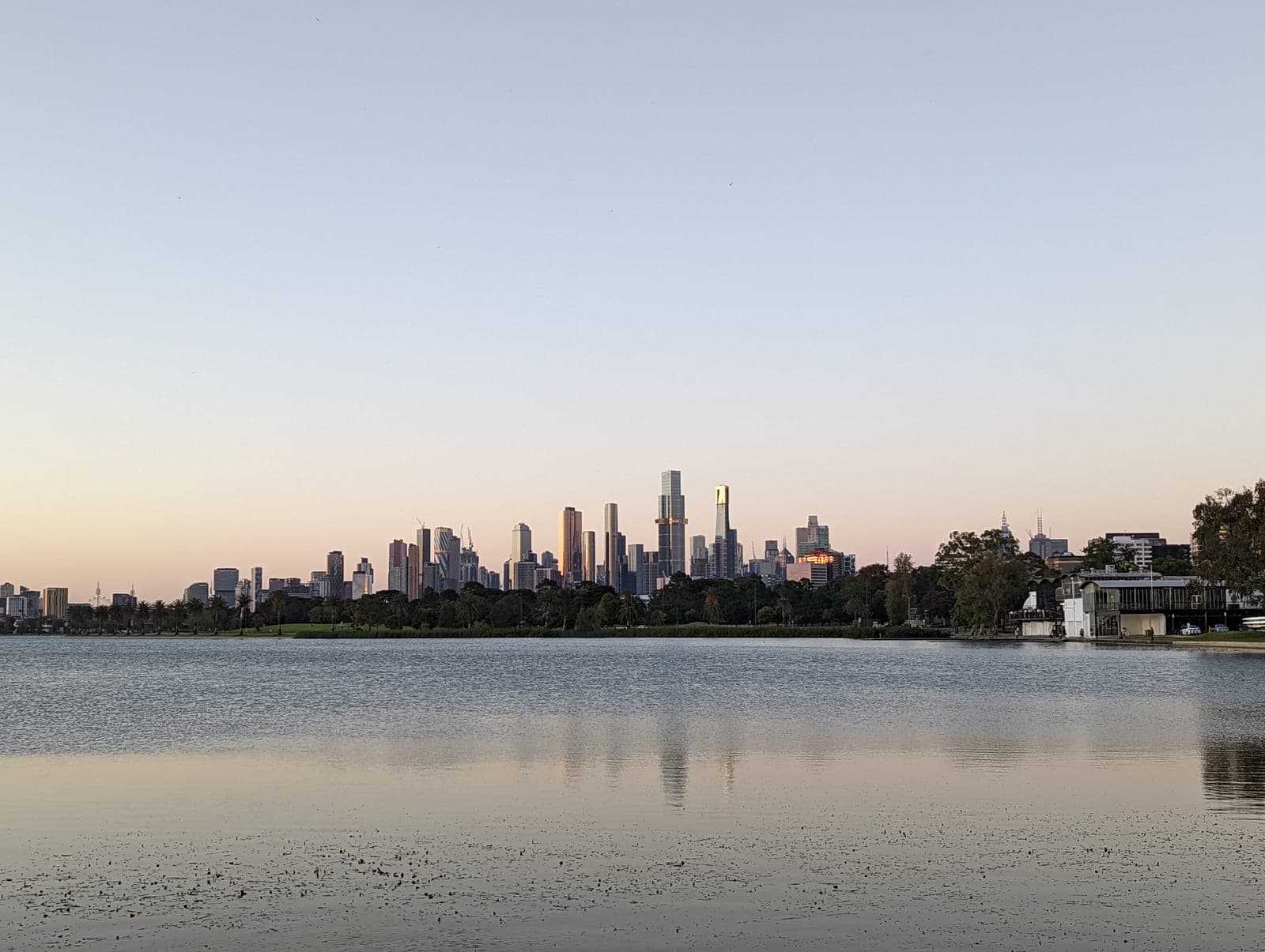 The Melbourne skyline seen across a lake at dusk, under a clear sky, with the water still and the towers catching the last of the light.