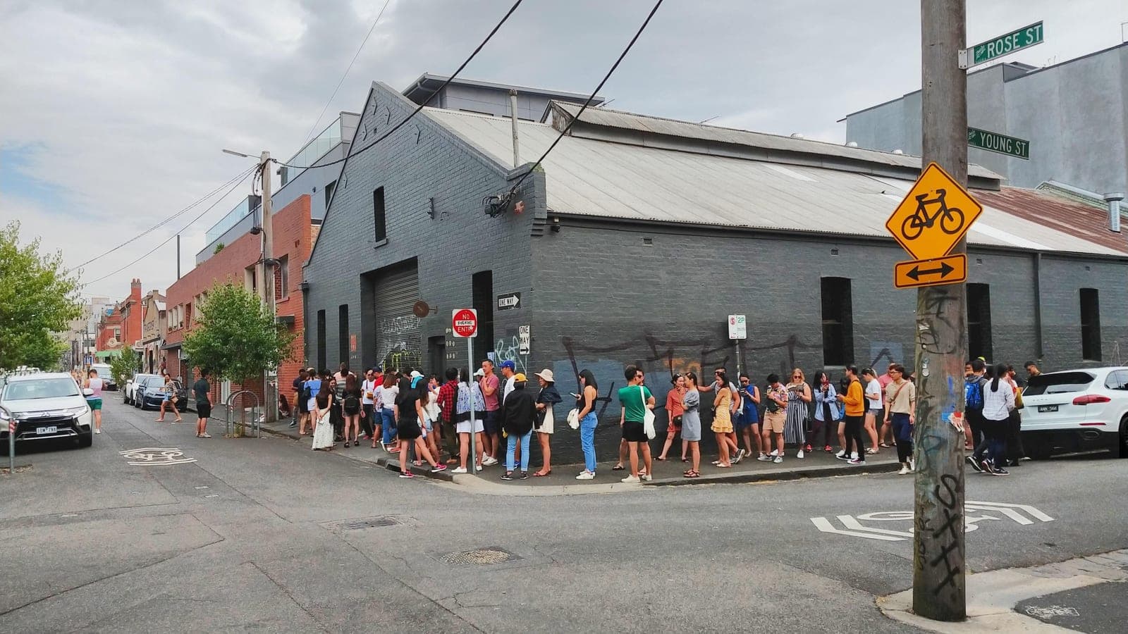 A long queue of people stretching along a footpath outside a blacked-out warehouse-style shopfront on a Fitzroy street corner, with no visible signage.