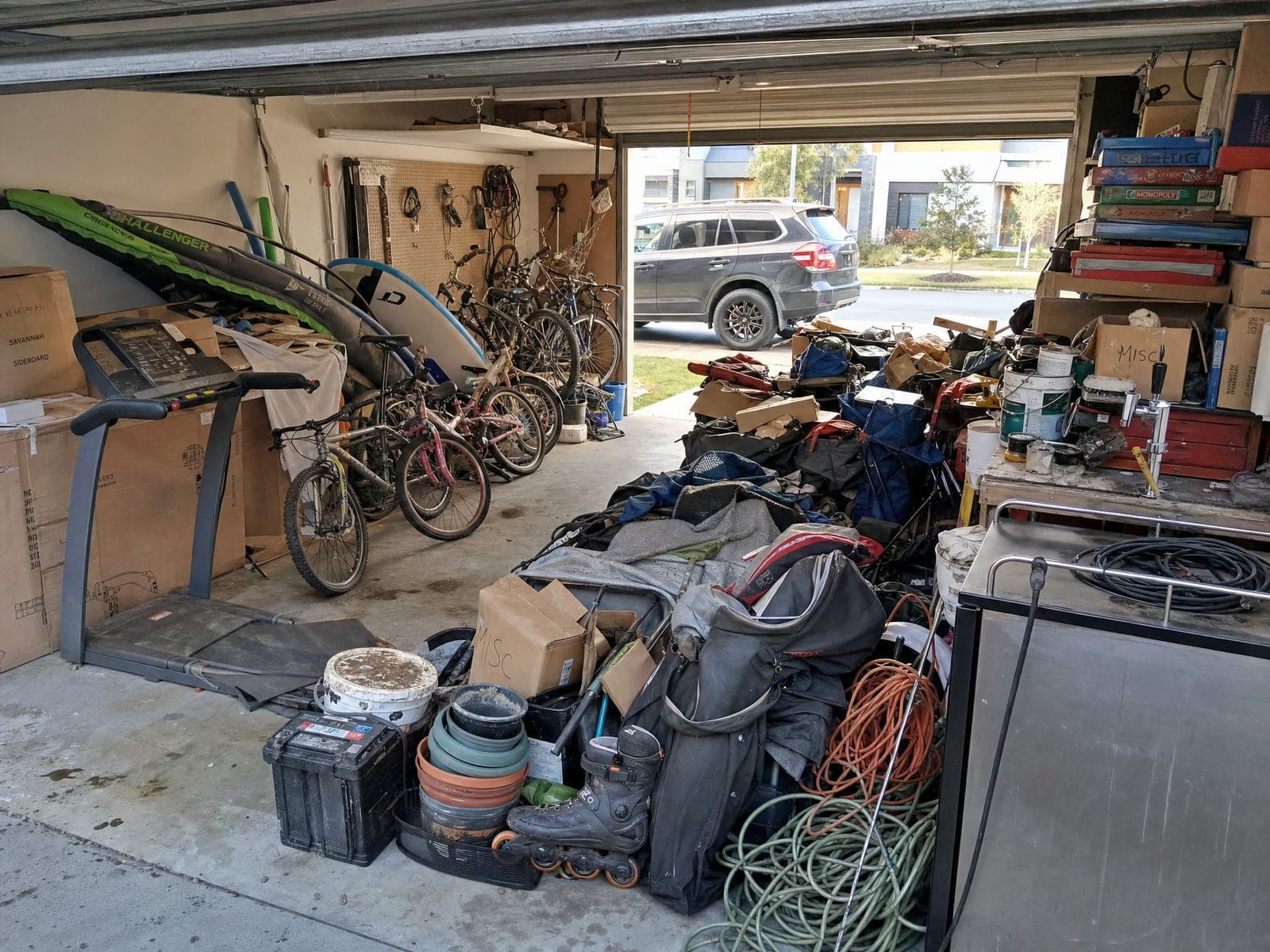 A double garage packed with a treadmill, surfboards, several bikes, boxes marked misc, paint tins and a fridge, with an SUV parked outside on the driveway.