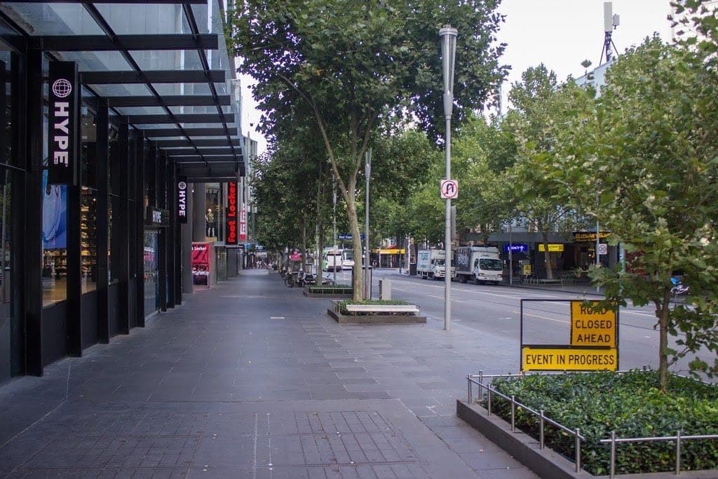 An empty city footpath and road lined with trees and shopfronts, with a yellow sign reading Road Closed Ahead, Event In Progress.