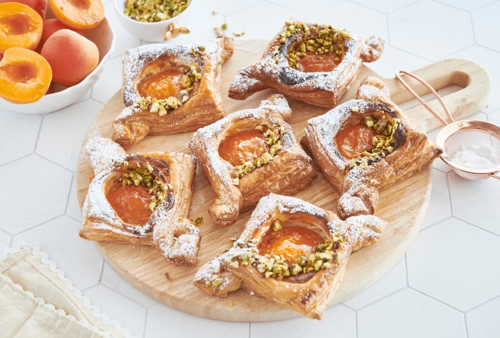Six square puff pastry danishes topped with apricot halves, crushed pistachio and icing sugar, arranged on a wooden board beside a bowl of fresh apricots and a copper sugar sifter