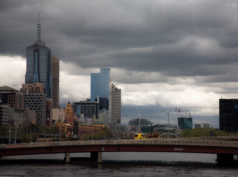 The Melbourne skyline under a heavy, overcast grey sky, seen across the Yarra River with a bridge crossing in the foreground.